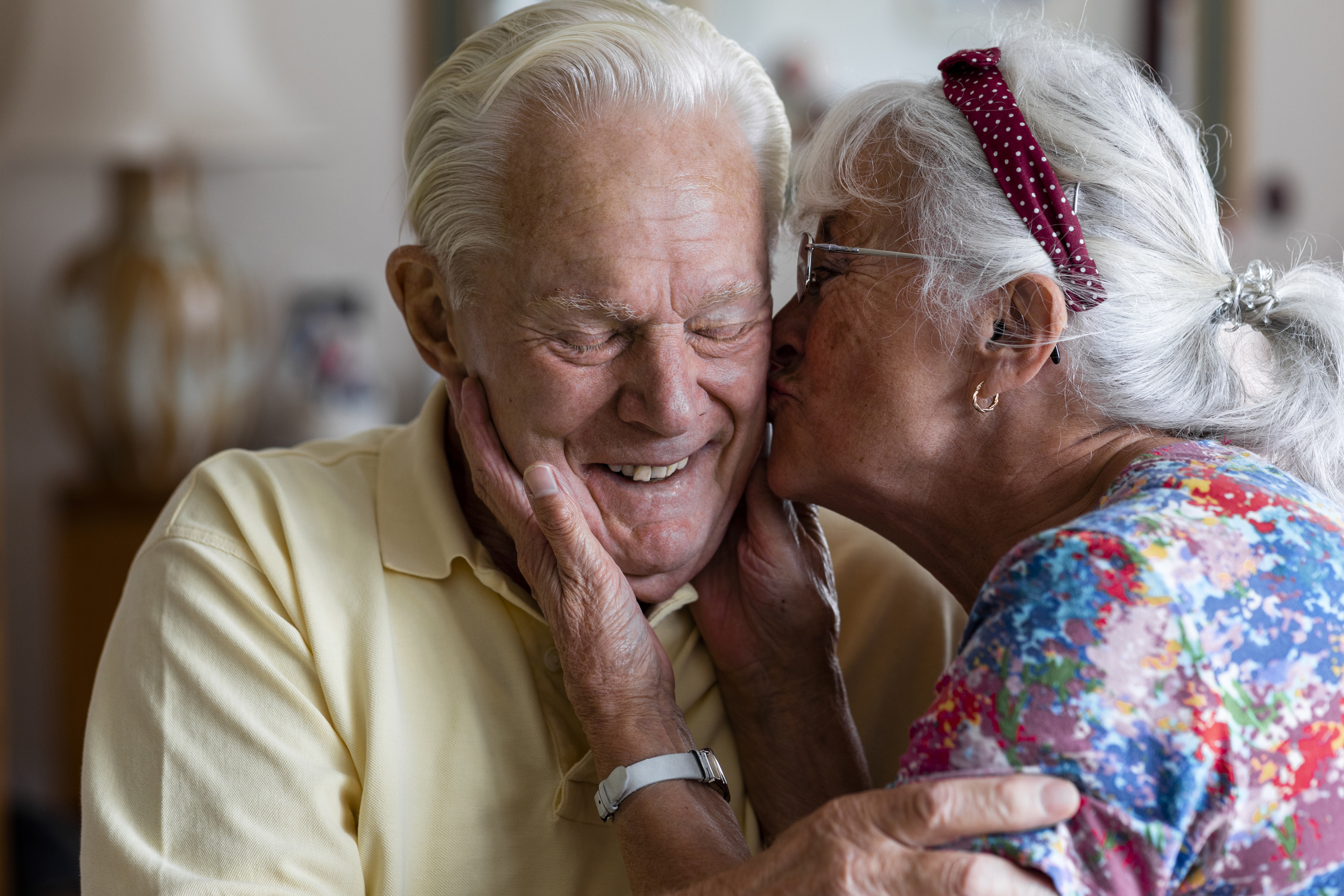Senior couple at home in the North East of England. The wife is kissing her husband's cheek while he laughs with his eyes closed.
